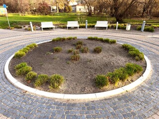 A calm park scene with a round garden bed and soft green shrubs framed by bright benches and warm...