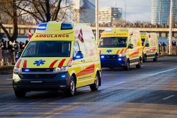 Series of yellow ambulances with active blue lights, Latvian flags and medical markings driving down multilane road as crowds gather behind roadside barriers