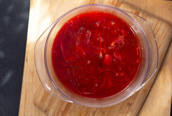 A clear glass container of homemade red borscht sits on a wooden cutting board, with rich beetroot broth and visible vegetables. Soft window light highlights the deep red tones and warm homemade feel 