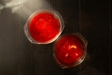 Two bowls of homemade red borscht are arranged diagonally on a dark wooden surface, with strong warm light illuminating the rich beetroot color and ingredients. The scene highlights the traditional
