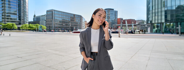 Young Asian successful businesswoman leader wearing suit standing in big city talking on mobile...
