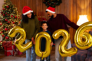 Joyful Latin family marks the start of 2026, holding large gold balloons in front of Christmas tree, posing in room with cozy holiday decorations