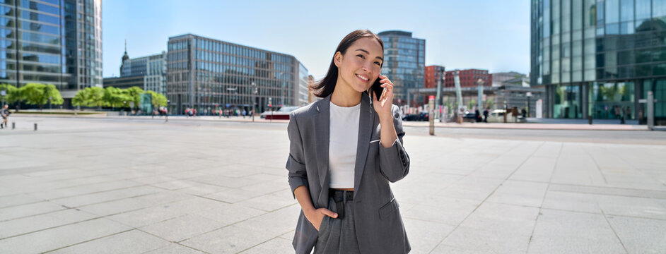 Young Asian successful businesswoman leader wearing suit standing in big city talking on mobile phone. Smiling woman making business call on cell walking on busy downtown street outdoors