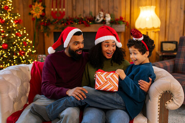 Excited Latin parents in Santa hats giving Xmas present to their son, family celebrating New Year, sitting on sofa and exchanging gifts
