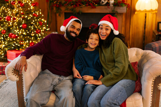 African American family of three sharing joyful moment in living room decorated for Christmas, wearing Santa hats and smiling, sitting on couch near beautifully lit Christmas tree - Powered by Adobe