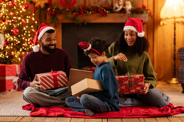 Surprised Latin boy opening gift box, sitting on carpet floor with parents in cozy living room decorated for Christmas, exchanging gifts