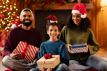 African American family exchanging Xmas gifts, boy and parents holding presents, smiling at camera, sitting near Christmas tree at home