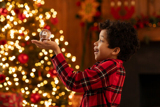Overjoyed black boy in red checkered shirt smiling as holding snow globe, admiring its shimmering details, beautifully decorated Christmas tree glowing with warm lights on background
