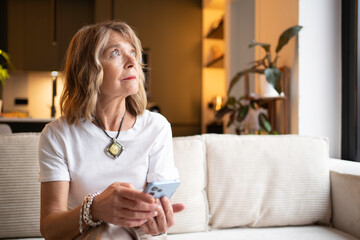 Senior woman sitting on couch holding phone, looking up with thoughtful expression