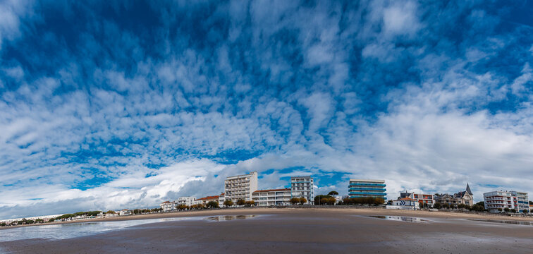 Panoramic shot of Royan Beach features a wide expanse of sand and calm water under a dramatic sky filled with scattered clouds. Modern buildings line the distant shore.