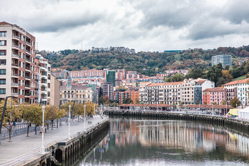 wide view of the Bilbao riverfront, with colorful buildings reflected in the calm water. The city rises up a wooded hillside under a dramatic, cloudy sky, showcasing urban and natural elements.