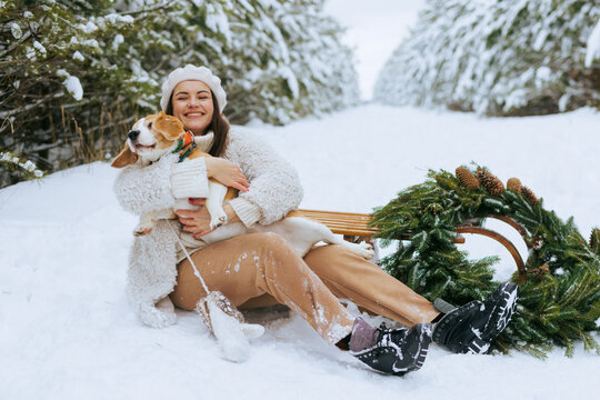 Smiling woman with dog on sled in snowy winter forest with Christmas wreath