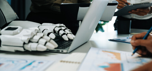 A modern workspace scene showing a robotic hand beside a laptop, with a business team engaged in project discussion, analyzing data, and fostering creativity.Noogenesis