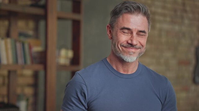 Portrait of happy confident casual middle aged man at home in loft room. Fit and healthy active senior male with grey hair looking at camera, smiling. Books in background, smart older guy.