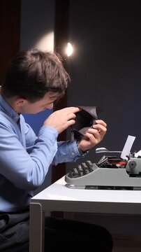 vertical video Young man in a blue shirt holds an empty wallet with a concerned face, sitting at a desk next to a vintage typewriter, representing financial hardship and the struggles of an author