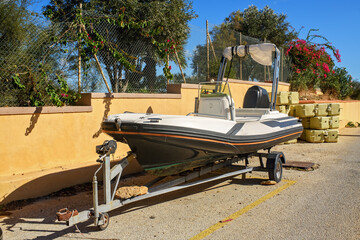 A road trailer with an inflatable motorboat is parked on the street against a wall, with flowering plants and modular pontoon blocks in the background.