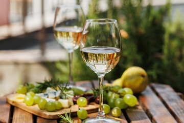 Elegant garden table with two glasses of white wine, green grapes, pear, and cheese board under sunlight
