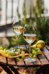 Rustic outdoor setup with two white wine glasses, grapes, cheeses, pear, and rosemary on garden table