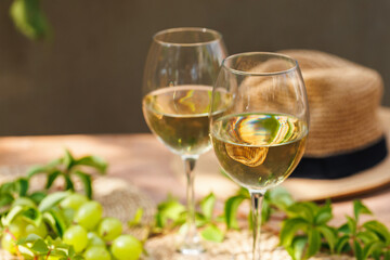 Two white wine glasses in sunlight with grapes and straw hat on wooden table with greenery..