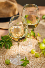 Two glasses of white wine with green grapes and straw hat in bright outdoor setting..