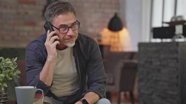 Happy middle aged man talking on phone call at home. Mid adult older male in glasses smiling, communicating. Telecommunication, mobile technology.