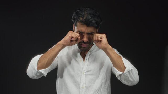 Young hispanic man rubbing his eyes in a black studio setting, wearing a white shirt and a tired expression; fatigue.