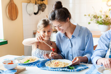 Happy family dinner. Mother and daughter eating pasta at home kitchen, enjoying healthy comfort food for children. Homemade ketchup, tomato sauce, mac and cheese. Candid, authentic lifestyle moment