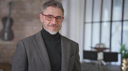 Portrait of happy casual middle aged man at home in industrial loft room. Smart looking businessman with grey hair looking at camera in glasses, jacket and turtleneck, smiling. Trustworthy, skilled.