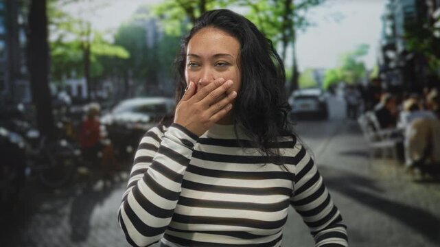 Woman hand on chest and hand to mouth on street in striped top, open mouth and raised eyebrow surprised expression; surprise wonder.