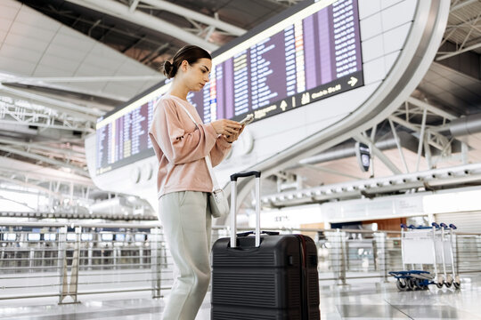 Young woman with suitcase stands in modern airport terminal, checking smartphone, surrounded by departure boards and spacious waiting area, capturing travel anticipation and excitement - Powered by Adobe