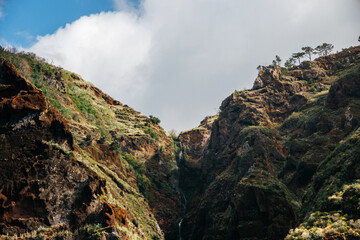 Majestic mountain landscape featuring steep cliffs, lush greenery, and a cascading waterfall, under a bright blue sky with fluffy clouds, showcasing natural beauty and tranquility