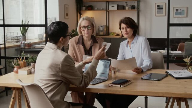 Two middle-aged Caucasian women in formal outfits having business discussion with their supervisor at desk in modern office