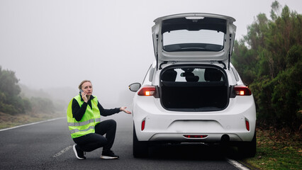 Female driver in reflective vest kneeling beside white car with open trunk on foggy road, demonstrating roadside assistance and vehicle troubleshooting in challenging weather