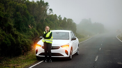 Caucasian woman in reflective vest stands beside a white car on a foggy roadside, showcasing roadside assistance and safety in a serene, natural environment