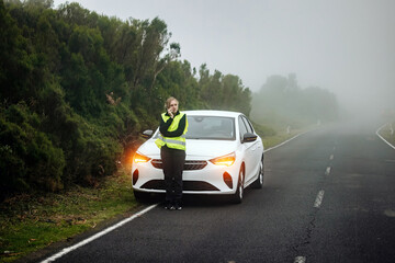 Female driver in reflective vest standing beside white car on foggy roadside, using phone for assistance, showcasing roadside emergency situation and safety awareness