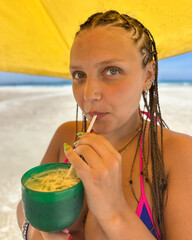 A young woman in a bikini with braided hair enjoys a  cocktail  in vibrant green glass  on a sunny beach.