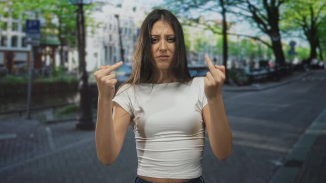 Woman holding up both middle fingers toward camera on a city street with visible bellybutton piercing and tense facial expression; defiance.