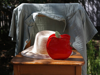 Red apple-shaped novelty bag and a straw hat on a wooden chair, with a shirt hanging on the backrest