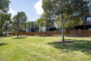 Green space with trees, a lawn with a row of modern townhouses in a suburban Melbourne with a timber boundary fence. Concept of medium-density housing, and well-maintained communal outdoor areas.