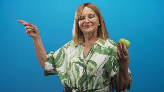 Woman senior points finger and holds green apple with hand visible, smiling in a blue studio backdrop wearing leaf patterned blouse; healthy choice confidence wellbeing.