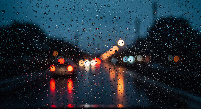 Driving through a city at night, the car windshield is covered in raindrops, creating a bokeh effect from the streetlights and traffic