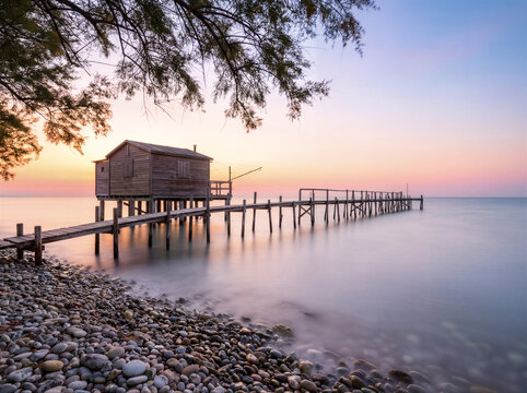 Wooden Fishing Hut On Pier At Sunset With Silky Sea And Rocky Beach