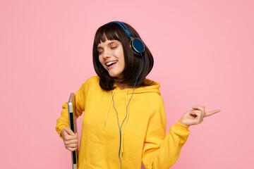 A cheerful young woman in a bright yellow hoodie dances with headphones on, holding a skateboard against a pink studio backdrop, capturing a playful, carefree mood and creative lifestyle.