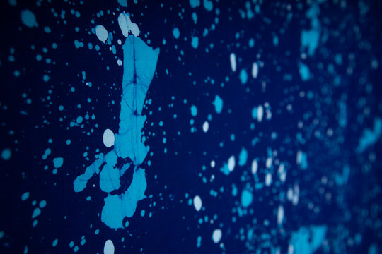 An angled close-up of a dark blue batik fabric, highlighting a large light blue splatter (ciprat) with subtle white specks, creating an abstract, textured depth