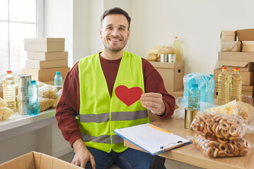 Portrait of smiling young man volunteer holding red heart in hands and looking at camera working at charity center. Male person volunteering in charitable foundation. Humanitarian aid concept.
