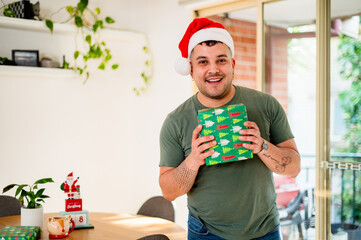 A joyful man celebrates Christmas at home, holding a wrapped gift while wearing a santa hat