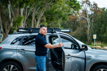 A man in his 30's smiles while opening his car door in a park setting.