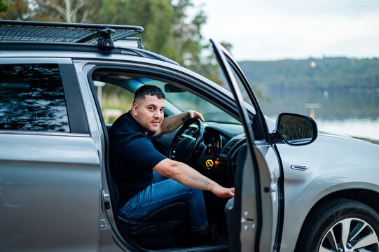 A man sits in the driver's seat of a parked car, taking in the fresh air at a park near the water
