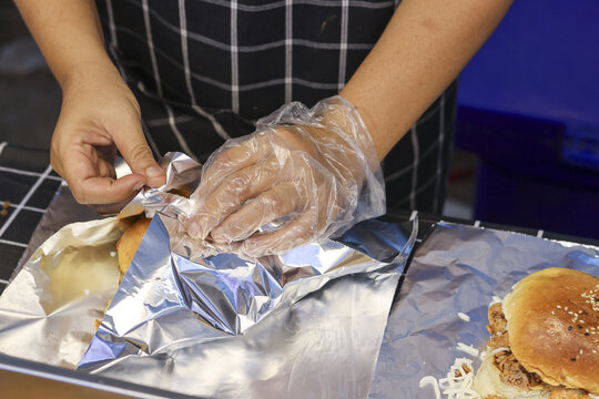 Careful chef wearing glove wraps fresh burger in foil for takeaway. Food preparation with focus on hygiene and service at restaurant or street food stall for delivery