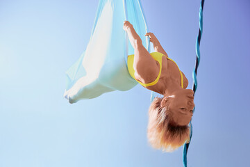 Slim mature woman keeps active and exercises her body through aerial yoga suspended upside down from a hammock